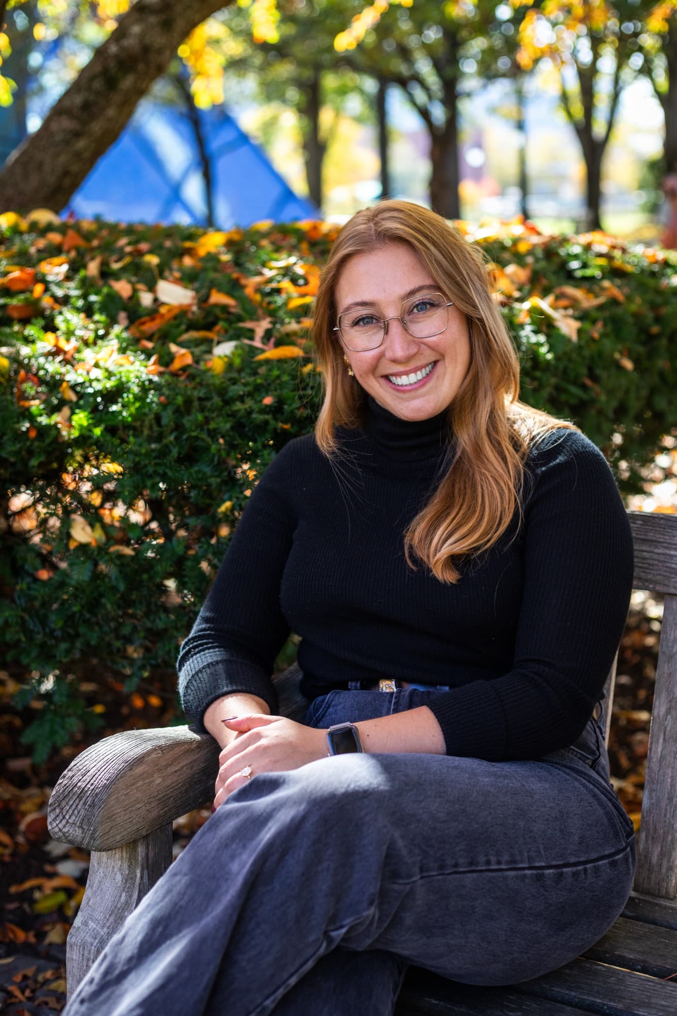 Portrait of a smiling woman with glasses seated on a bench among autumn leaves